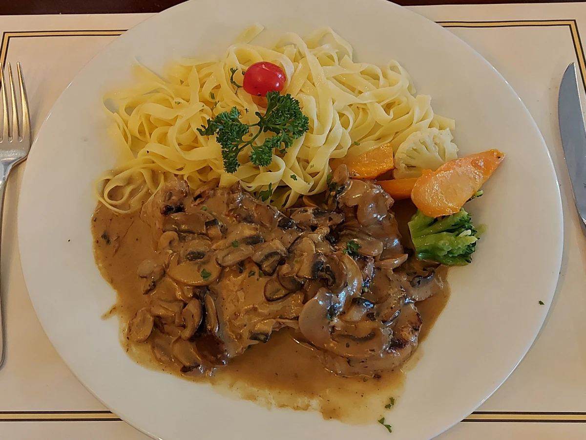 Zwei Schweinsschnitzel mit frischer Pilz-Rahmsauce, serviert mit Tagliatelle, begleitet von Blumenkohl, Karotten und Broccoli, garniert mit Petersilie und einer halben Cherrytomate, auf einem weissen Teller. Ein appetitliches Mittagsgericht.