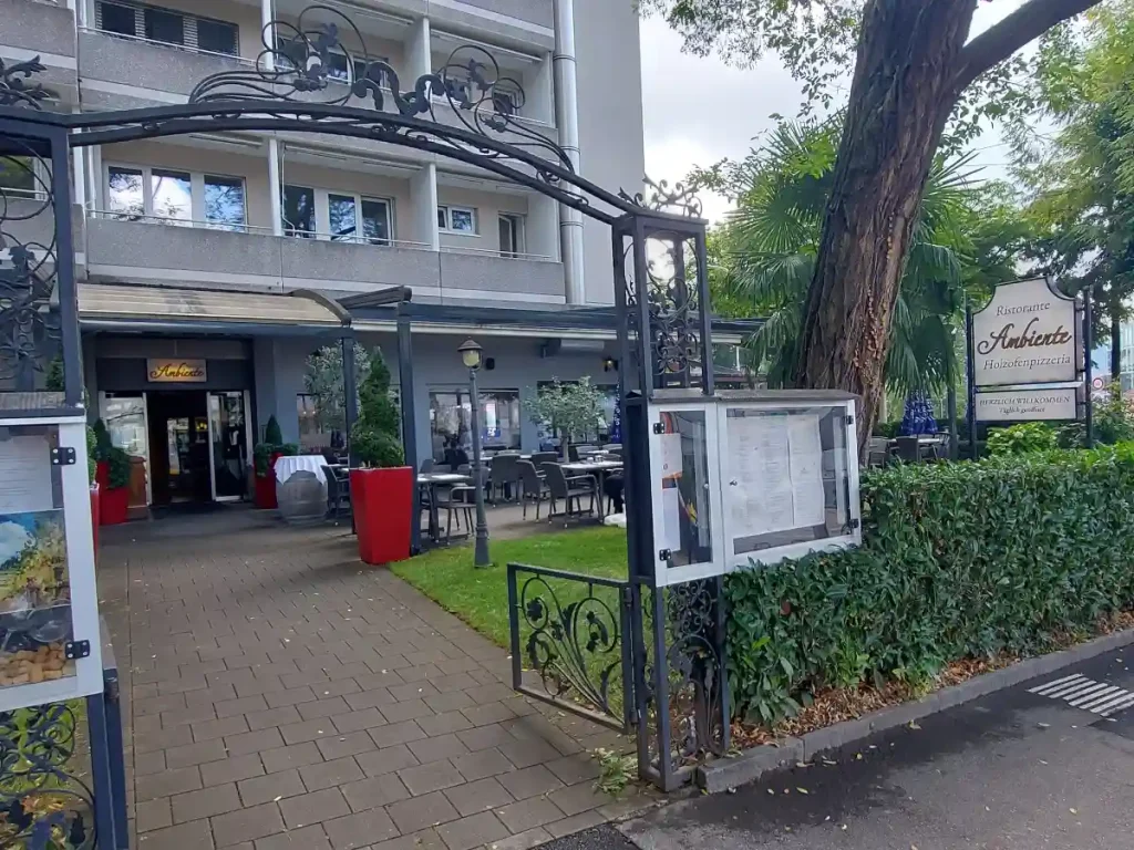 Der Eingangsbereich des Ambiente Bern von aussen: Ein schmiedeisernes Tor mit beidseitigen Speisekarten-Schaukästen führt zu einem durch eine Hecke abgegrenzten Vorgarten mit Rasen, einer möblierten Terrasse und der Eingangstür des Restaurants.