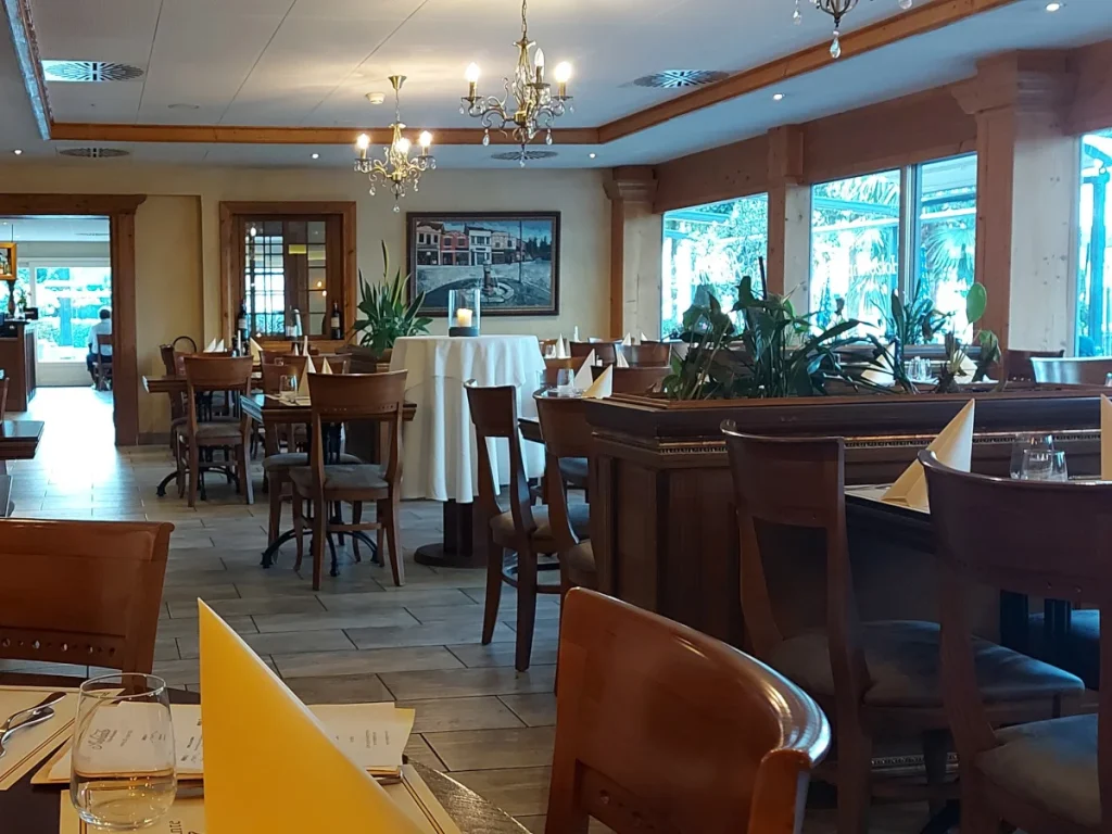 Inneneinrichtung des Ambiente Bern: Ein heller Raum mit vielen Fenstern, dunklen Holztischen, die mit hochwertigen Papierservietten gedeckt sind, und passenden dunklen Holzstühlen mit Sitzpolstern. Die Wände sind mit Bildern italienischer Motive dekoriert.