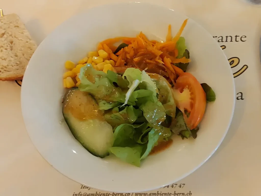 Kleiner Menüsalat auf einem weissen, tiefen Teller, mit grünen Salatblättern, einem Stück Tomate, Karotten, Mais und einem Stück Gurke, übergossen mit italienischer Salatsauce.