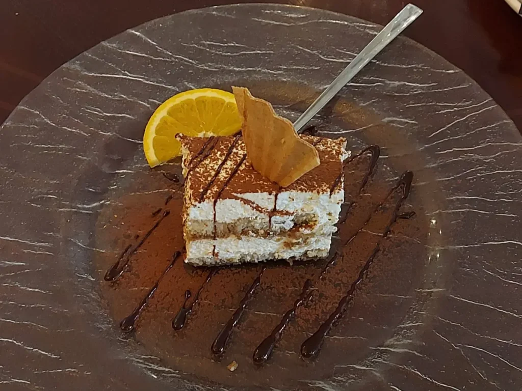 Ein quaderförmiges Stück Tiramisu, garniert mit einer Kekswaffel, serviert auf einem runden, strukturierten Glasteller, dekoriert mit Kakaopulver, Schokoladensauce-Streifen und einer halben Orangenscheibe.