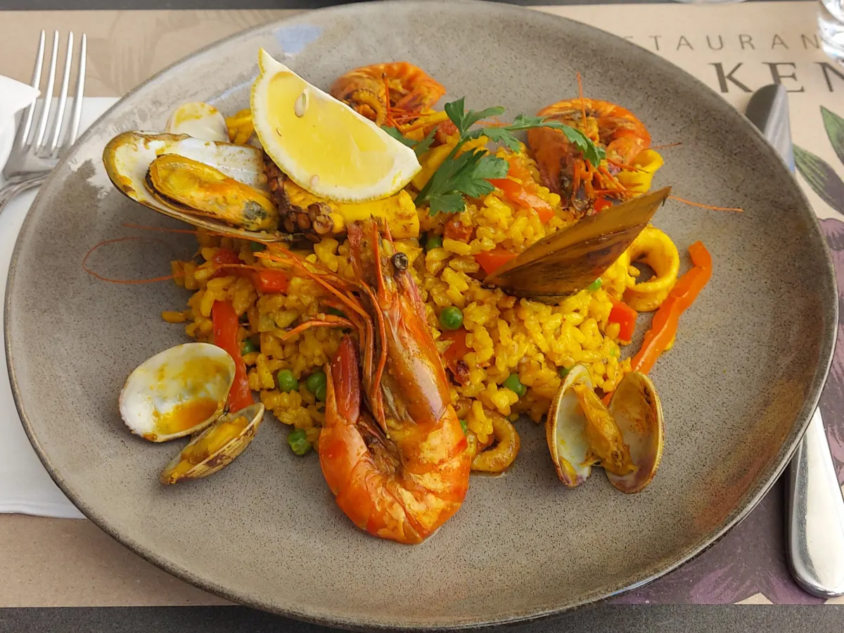 Nahaufnahme einer Paella des Falken Bern mit safrangelbem Reis, Riesencrevetten, Muscheln, Chorizo, Paprika und Zitrone auf einem grauen Teller.