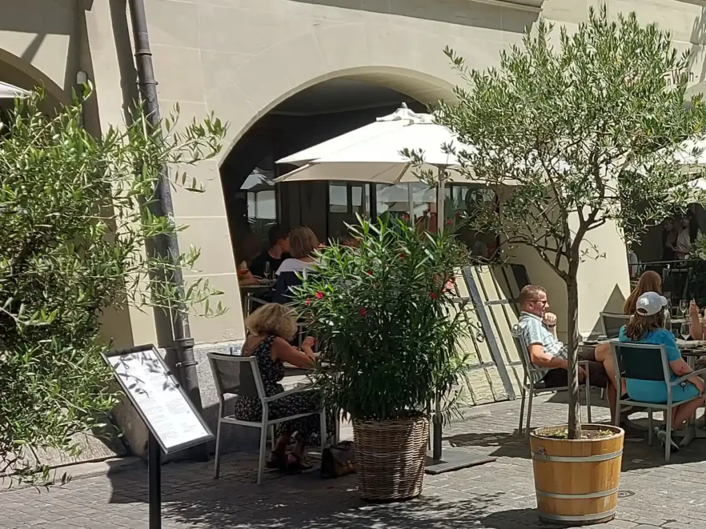 Terrasse des Falken Bern in der Berner Altstadt mit Gästen, üppiger Begrünung und weissen Sonnenschirmen.