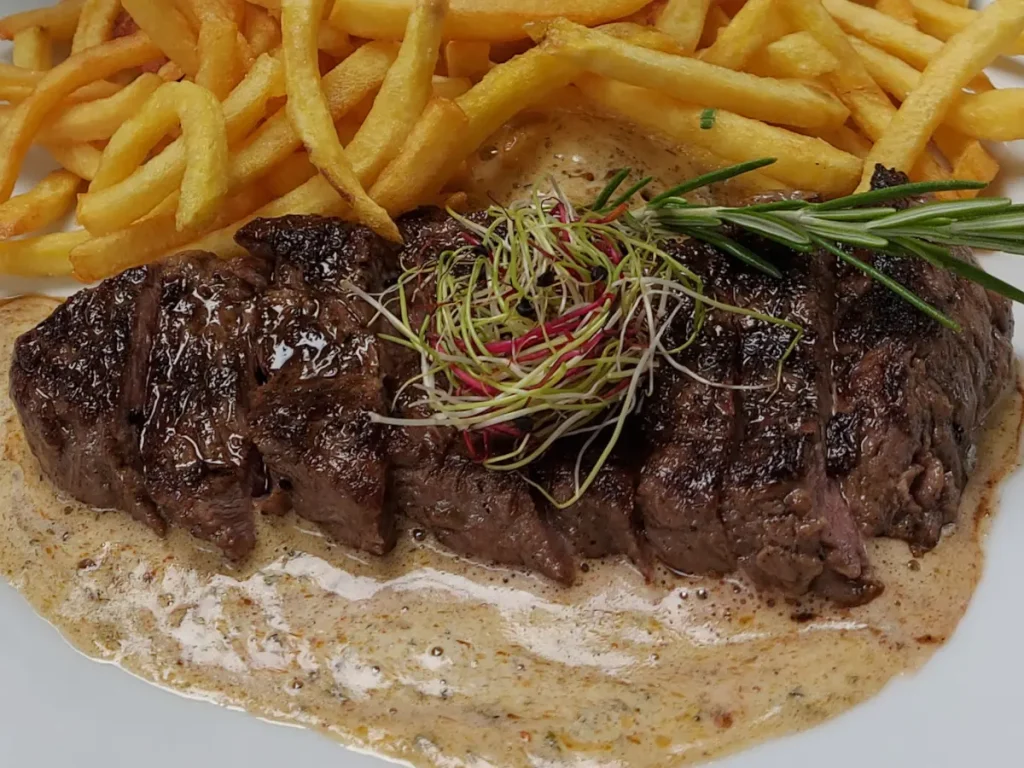 Nahaufnahme des saftigen, geschnittenen Entrecôte mit glänzender Café de Paris Sauce und danebenliegenden knusprigen Pommes Allumettes.