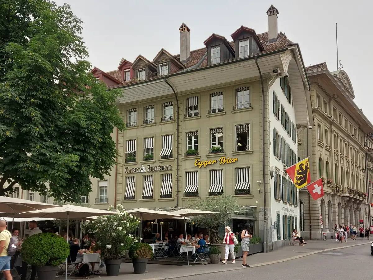 Die einladende Frontansicht des Restaurants Federal Bern, ehemals Café Fédéral, mit sonniger Terrasse, beigen Sonnenschirmen und einem charakteristischen Kastanienbaum am Bärenplatz.