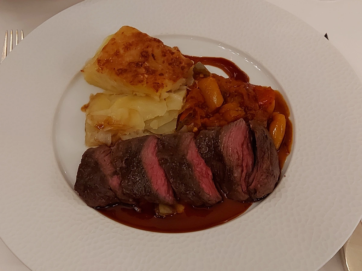 Ein zartrosa gegartes Flank Steak mit kross gebratener, goldbrauner Kruste liegt auf einer dunklen braunen Sauce in einem weissen Porzellanteller. Daneben befinden sich mediterranes Ratatouille – erkennbar durch gelbe Paprikascheiben – sowie ein viereckiges Stück Kartoffelgratin mit goldbrauner Gratinierung. Das Gericht wirkt klassisch elegant und entspricht dem gehobenen Stil der Jack's Brasserie im Hotel Schweizerhof Bern.