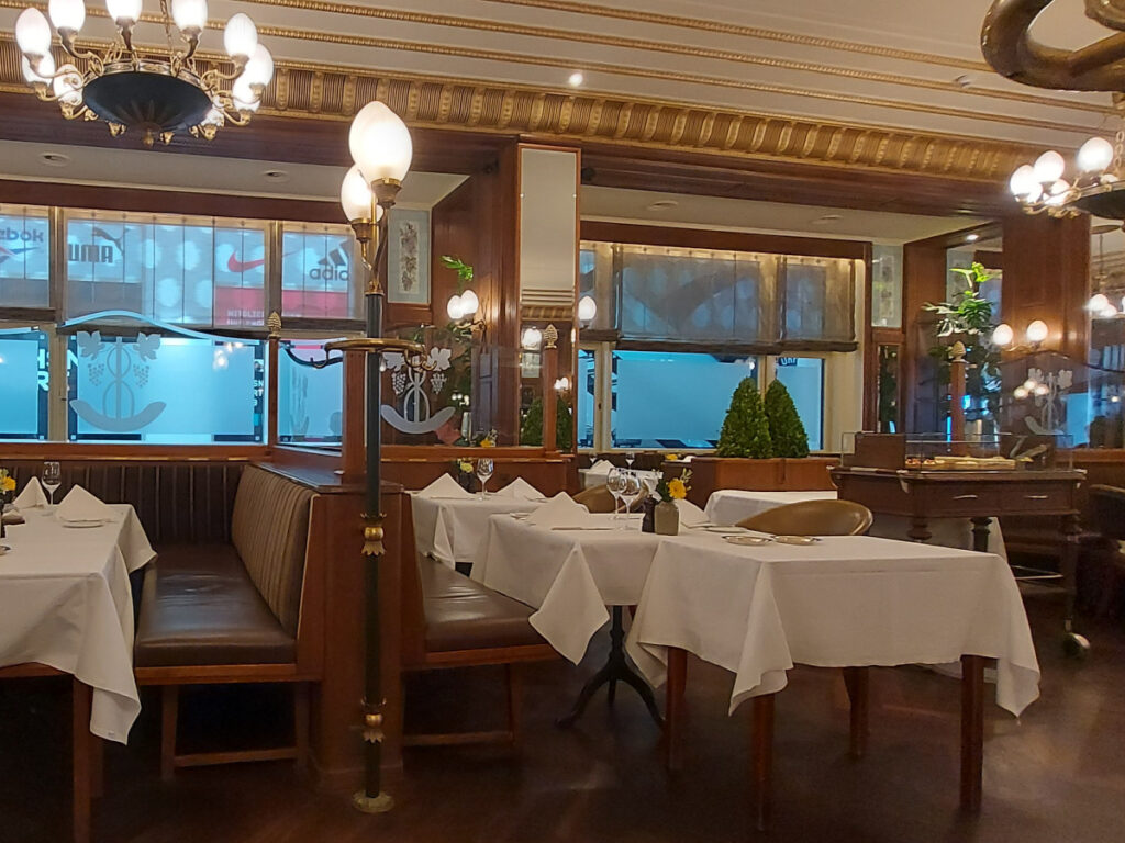 Das klassische, ehrwürdige Interieur der Jack’s Brasserie im Hotel Schweizerhof Bern – grosse Kronleuchter, grosse Fenster, Holztische mit weissen Tischdecken und gepolsterte Sitzbänke/Stühle.
