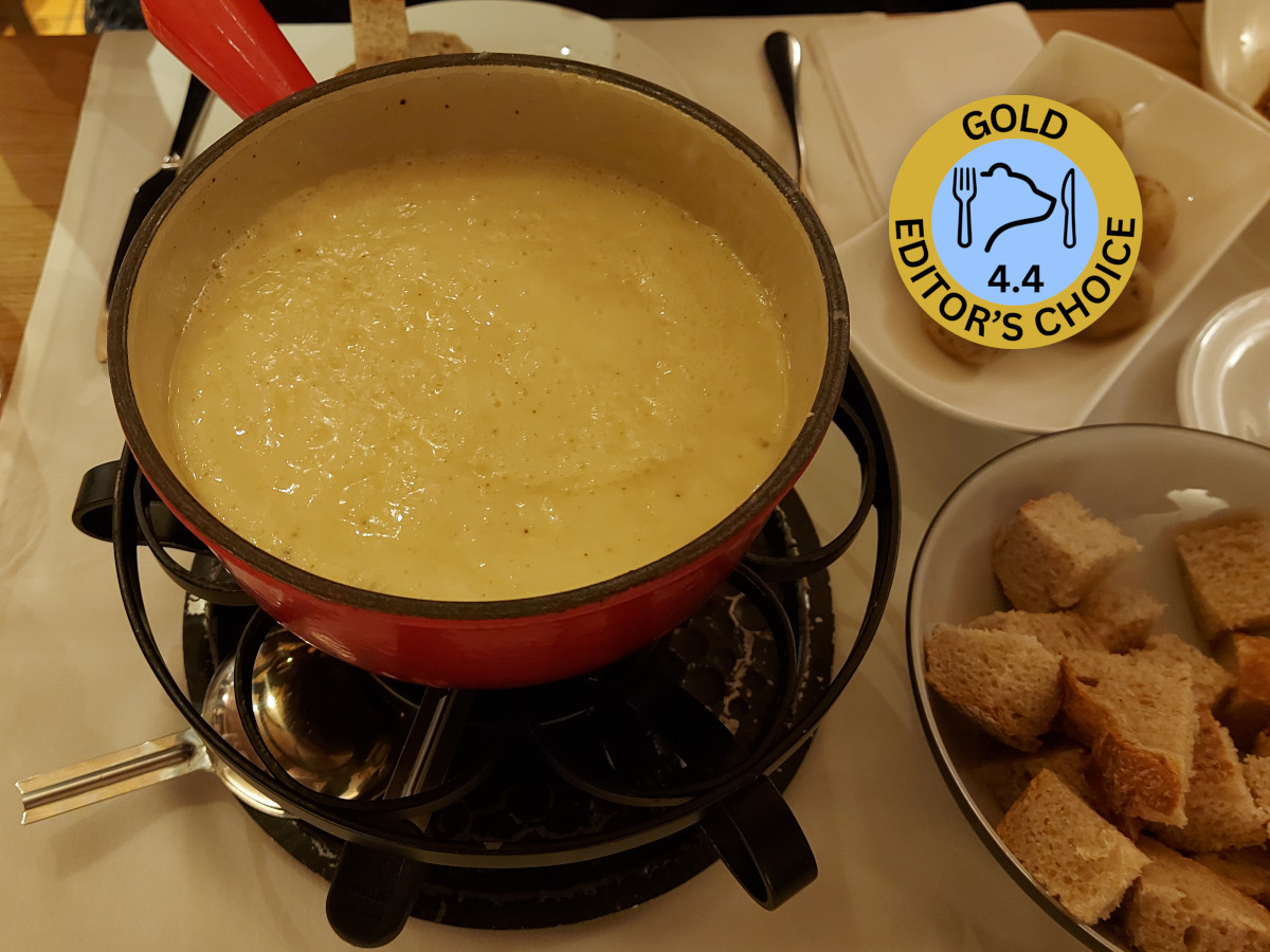 Käsefondue im roten Caquelon auf einem schwarzen Rechaud, ergänzt durch die goldene „Editor’s Choice“ Medaille von eateryfinds.com mit der Note 4.4.