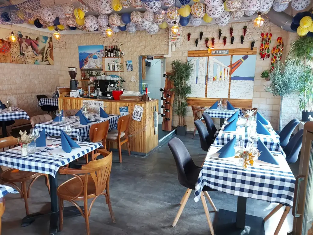 Innenansicht der Little Greek Taverna: Tische mit blau‑weissen Tischtüchern, mediterranem Dekor, Papier‑Ballons an der Decke.