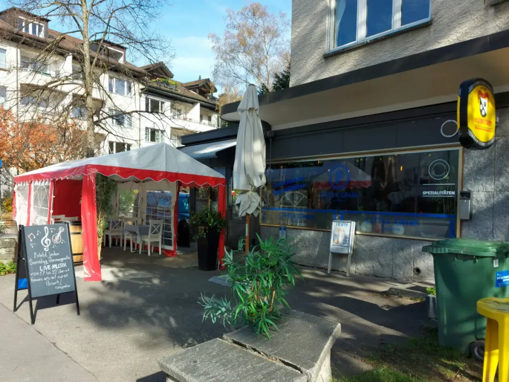 Aussenansicht der Little Greek Taverna in einem unscheinbaren beigen Gebäude mit großer Fensterscheibe, Restaurant‑Schriftzug, Vorzelt und Schild.