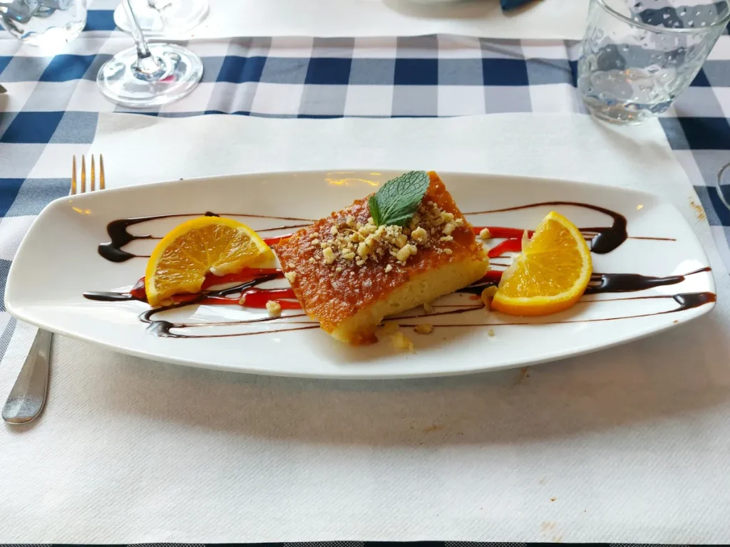 Griechischer Griesskuchen auf schön angerichtetem Teller, garniert mit Nüssen, Minzblatt und zwei halben Orangenscheiben; saftig‑feucht.