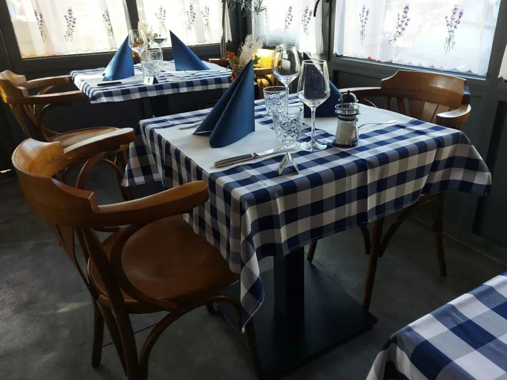 Nahaufnahme der Tische der Little Greek Taverna: blau‑weisse Tischtücher, weisse Unterlagen, blaue Servietten, Besteck und Gläser; heller Raum.
