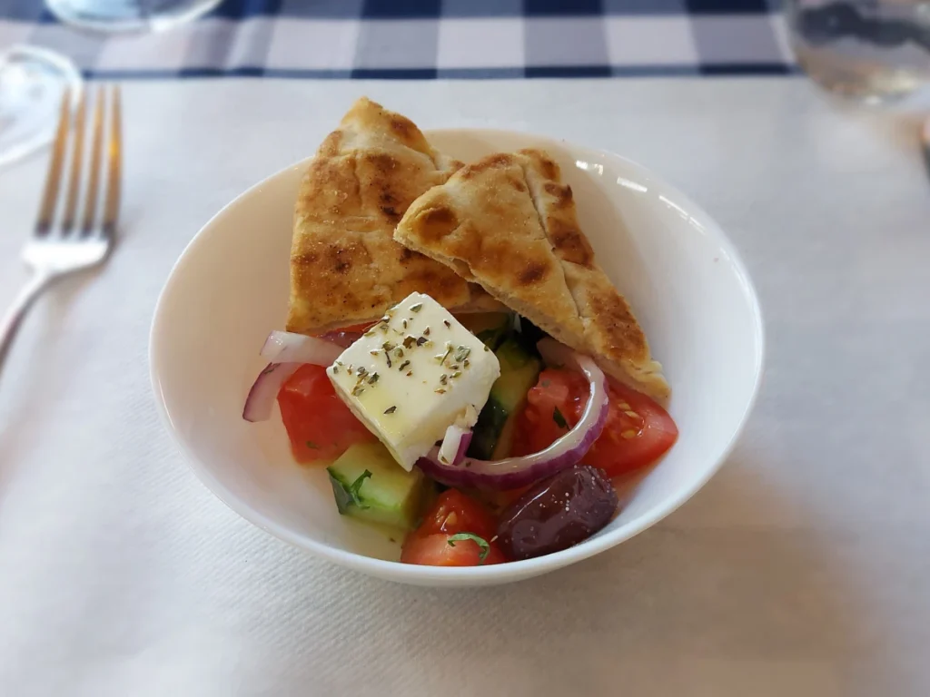 Kleiner griechischer Salat in weisser Schüssel: Tomaten, Gurken, violette Oliven, Feta und zwei Pita‑Brote.