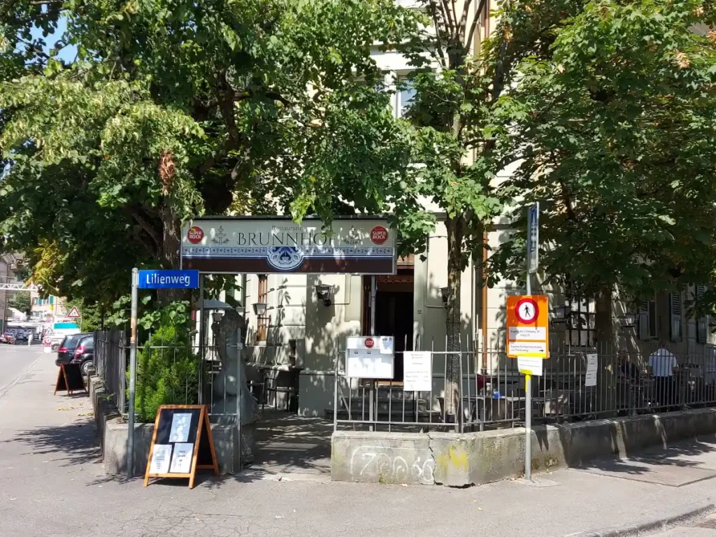 Aussenansicht des Restaurant Brunnhof in Bern: ein älteres Eckgebäude mit einer einladenden Terrasse im Schatten grosser Kastanienbäume, umgeben von einem schmiedeeisernen Zaun und einem "Brunnhof"-Schild.