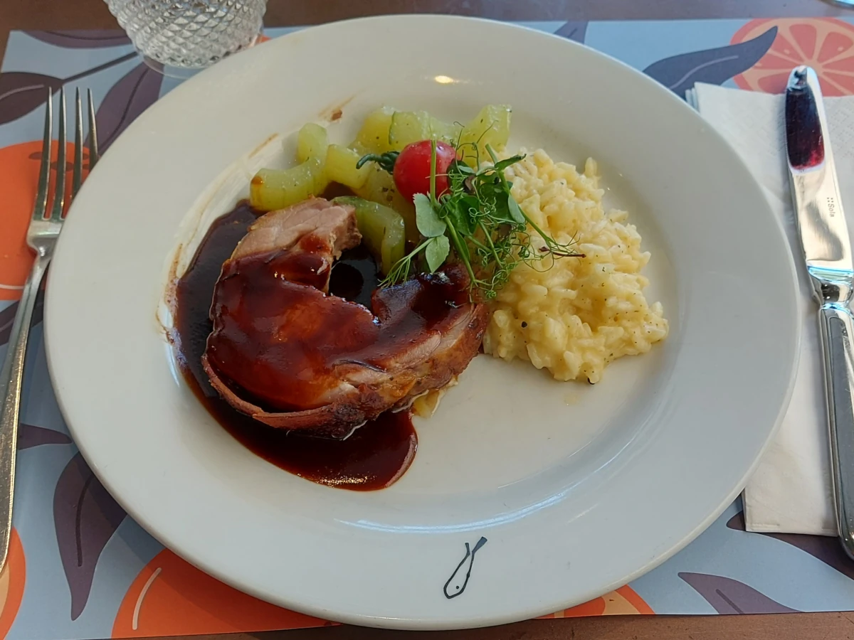 Spanferkel mit knuspriger Kruste, dunklen Fonds, cremigem Risotto und Gurkengemüse auf weissem Teller, umgeben von Besteck und Serviette, auf einem Tisch mit hellblau-orangem Tischtuch.