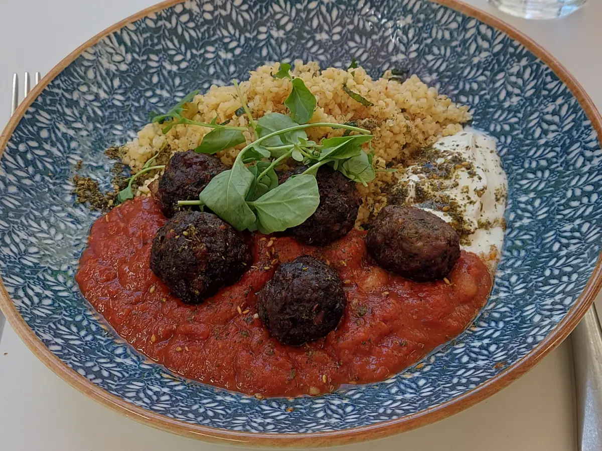 Dunkelbraun gebratene Rindshackfleischbällchen (Keftedes) in sämig-würziger Tomatensauce mit weissen Bohnen (Loubia), neben beigem Tabouleh und crèmigen Joghurt, garniert mit grünen Blättchen, serviert im Restaurant Sua Bern.