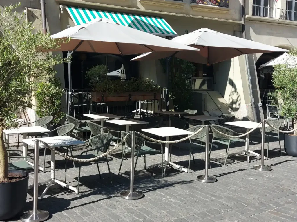 Aussenansicht der Terrasse des Restaurants Sua Bern in der Berner Altstadt: Die Lauben sowie pyramidenförmige Sonnenschirme spenden Schatten über weissen Gartentischen und -stühlen. Die Terrasse, welche sich auf Kopfsteinpflaster befindet, ist durch Seile zwischen hüfthohen Ständern von der Strasse abgegrenzt.